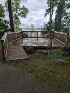 Scenic observation deck with cloudy skies over Cass Lake in the background. A gently sloped ramp comes down from the deck on the left side, but does not connect to the concrete path, leaving a 3" step onto grass and another step up onto the trail. 